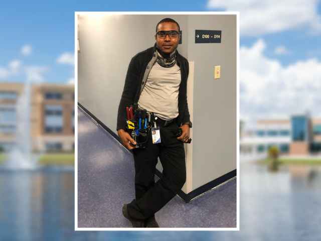 Fludumi Dade leans against a wall. He wears a tool belt.