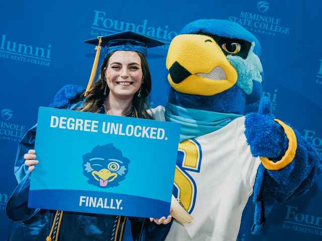 A graduate stands with Rally Raider and a sign that says "Degree Unlocked. Finally"