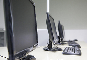Three black computer desktop monitors on a table in a classroom.
