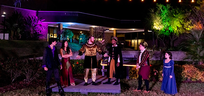Group of performers in Renaissance costumes on an outdoor stage at night with colorful lighting behind them.