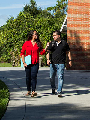 Two students walking together on a campus sidewalk, talking and smiling while carrying books.