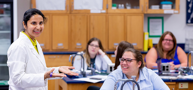 Instructor speaking to students in a science lab classroom, with students seated at lab tables listening and taking notes.