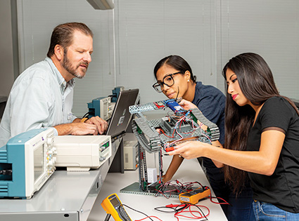 Students working with an instructor on an electronics project, assembling components and reviewing equipment at a lab workstation.