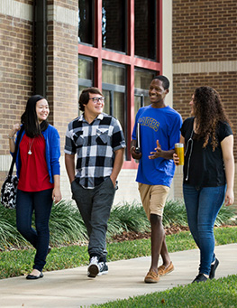 Four students walking along a campus sidewalk, talking and smiling as they pass a brick building.