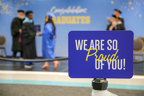 A sign that reads 'we are so proud of you' in white and yellow font on a royal blue background and a blurred image of a graduation state with students and professors behind the sign and one professor is holding a baby