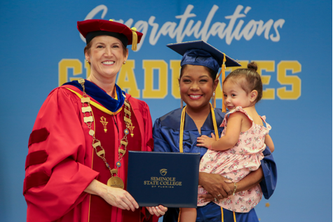 Dr Lorenz wearing a red graduation robe is holding a Seminole State College blue diploma with a lady student wearing a blue graduation robe that is also holding her toddler-aged daughter