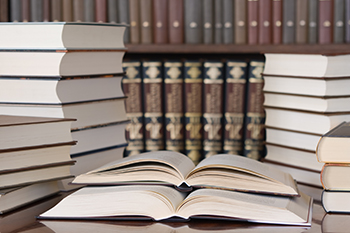 A pile of opened books on a table in a library with a bookshelf of books in the background