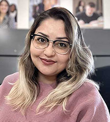 Person wearing a pink sweater seated indoors with photos on the wall behind them.