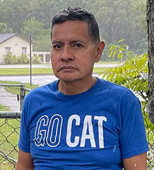 Person wearing a blue ‘GO CAT’ T‑shirt standing outdoors near a fence and trees