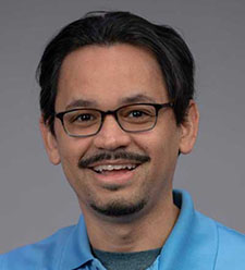 Person wearing a light blue collared shirt against a plain gray background.