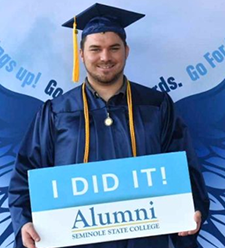 Graduate in a navy cap and gown holding an ‘I DID IT! Alumni – Seminole State College’ sign in front of a wing-themed backdrop.