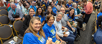 Group of employees sitting in the Sanford/Lake Mary C building smiling at the camera and waiting for the President's Address to start at Convocation.