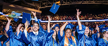 Group of SSC graduates, wearing blue graduation robes and tossing their hats into the air at the graduation ceremony inside UCF arena